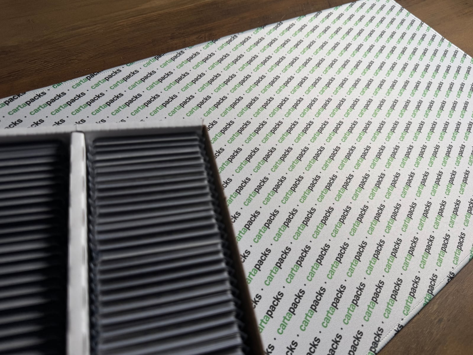 An open storage box of toploadered cards next to a cartapacks-branded playmat on a wood desk.